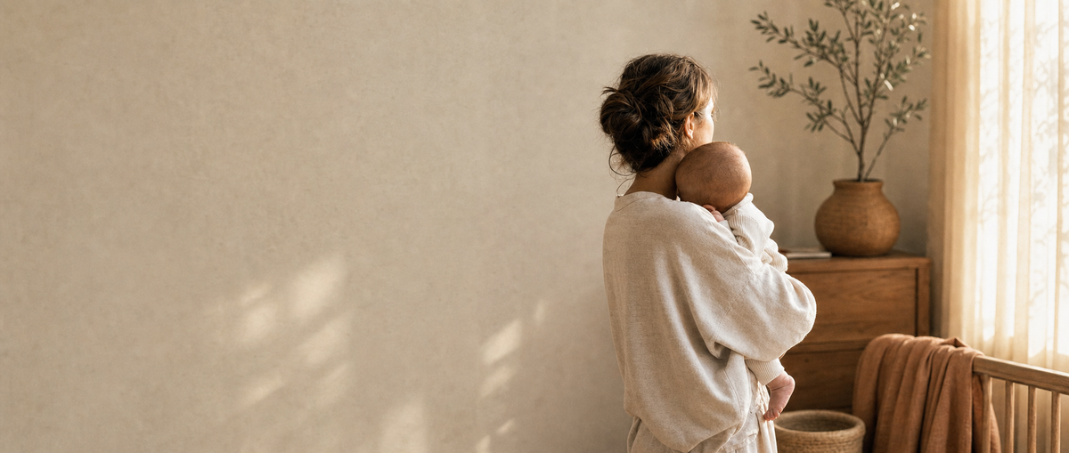 Motherhood collection banner: mother holding baby by a sunlit window with olive branch, soft editorial light